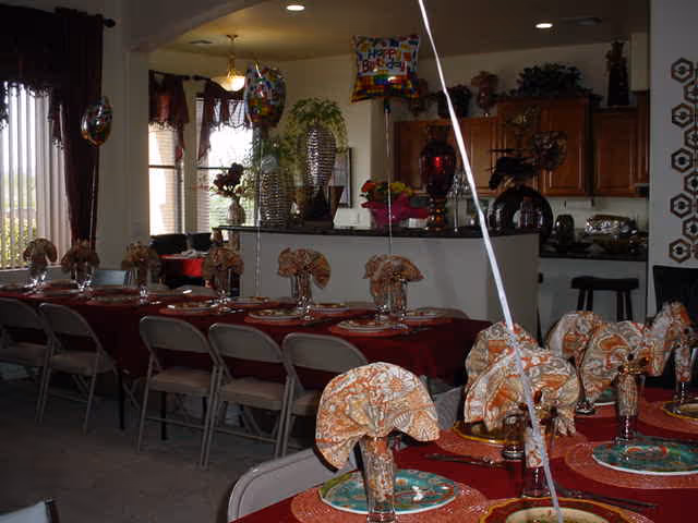 Interior view of a dining area in an assisted living facility decorated for a celebration with balloons and neatly arranged tables covered with red tablecloths, plates, and folded napkins. The background shows a kitchen area with wooden cabinets and bar stools.
