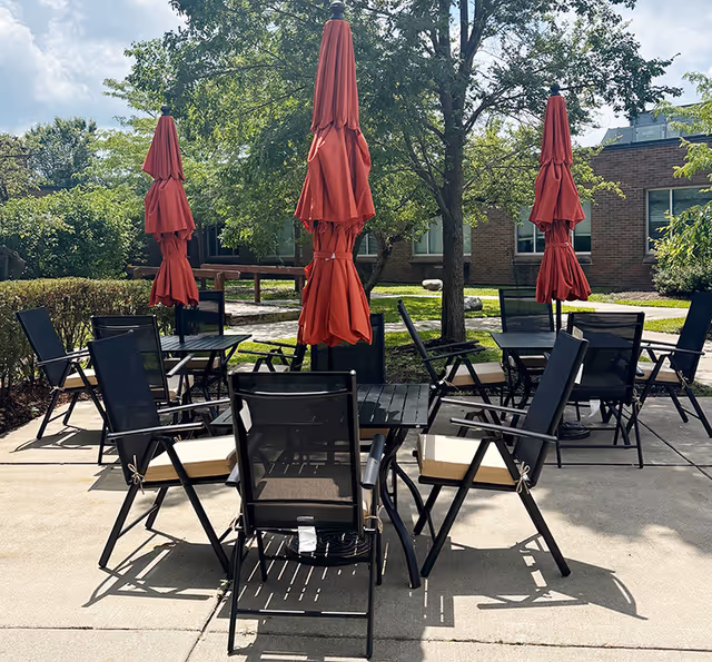 Outdoor patio with black metal tables and chairs, red umbrellas, near a brick building.