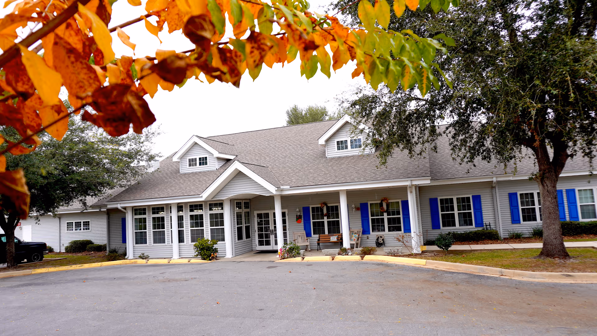 Front exterior view of a single-story senior living facility building with gray siding, blue shutters, and a covered porch with benches and rocking chairs. Trees with autumn leaves frame the top of the image.