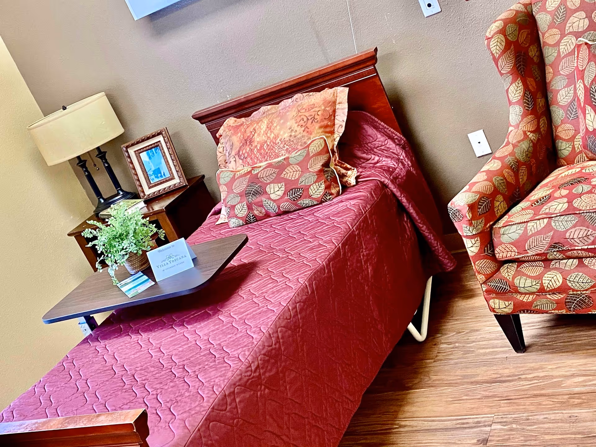 A burgundy quilted bed with matching pillows, a wooden nightstand, lamp, and patterned chair.