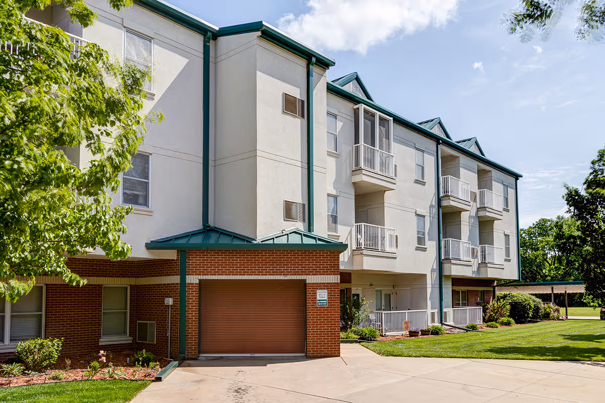 Exterior view of a three-story senior living facility building with white walls and green roof accents. The building features small balconies and a garage door on the ground level. There is a well-maintained lawn and trees surrounding the building under a partly cloudy sky.