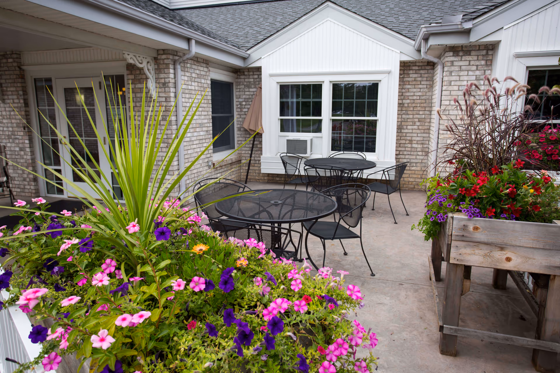 Outdoor patio with round metal tables, chairs, and bright flower planters by the building.