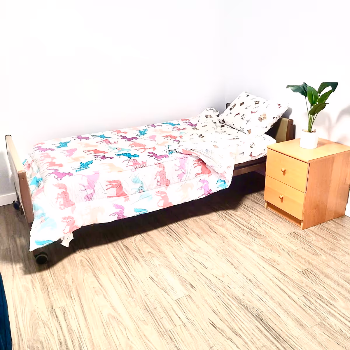 A single bed with colorful dinosaur-pattern bedding beside a wooden nightstand with a plant.