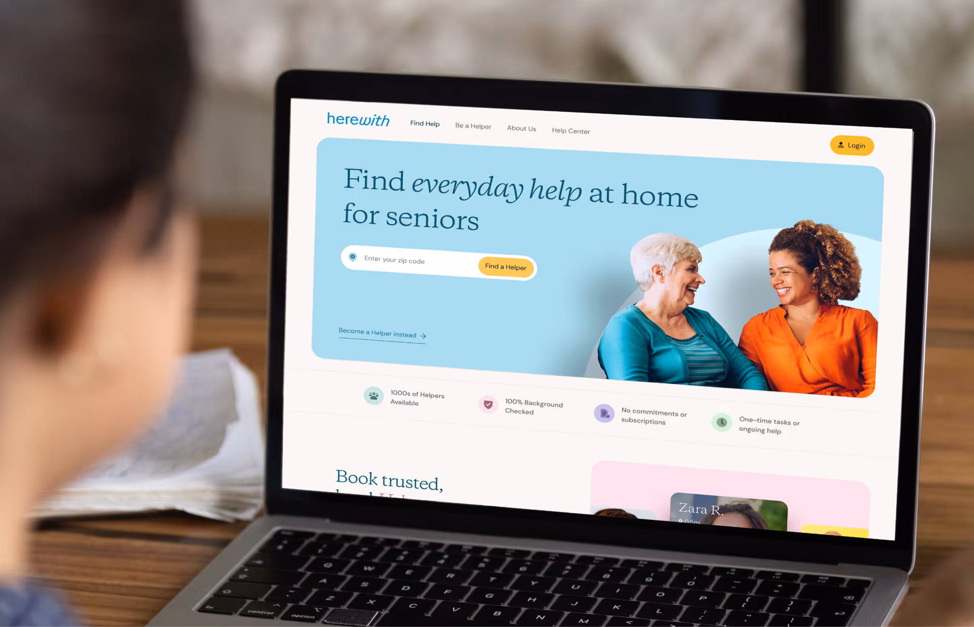 A person sits at a desk looking at a laptop displaying Herewith's senior care website.