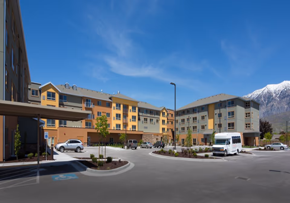 Exterior view of a multi-story senior living facility with a parking lot in front, several parked cars, and a shuttle van. The building is painted in shades of yellow and gray, with a clear blue sky and snow-capped mountains in the background.