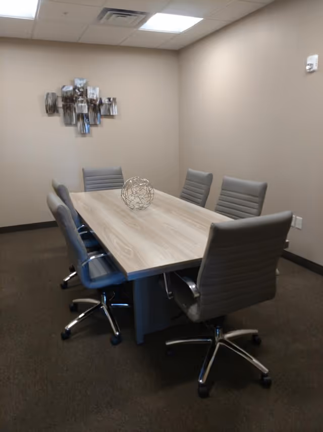 Small conference room with a rectangular light wood table surrounded by six gray swivel chairs and a decorative metal orb on the table with wall art on a neutral wall.