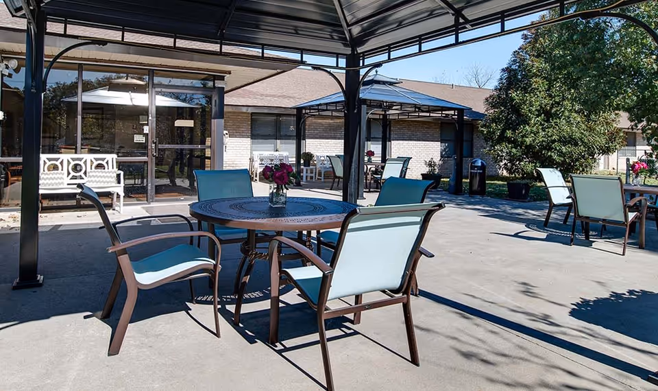 Outdoor patio area with multiple round tables and chairs under metal pergolas. Each table has a small vase with flowers. The patio is adjacent to a brick building with large glass doors and windows. Trees and greenery are visible in the background.