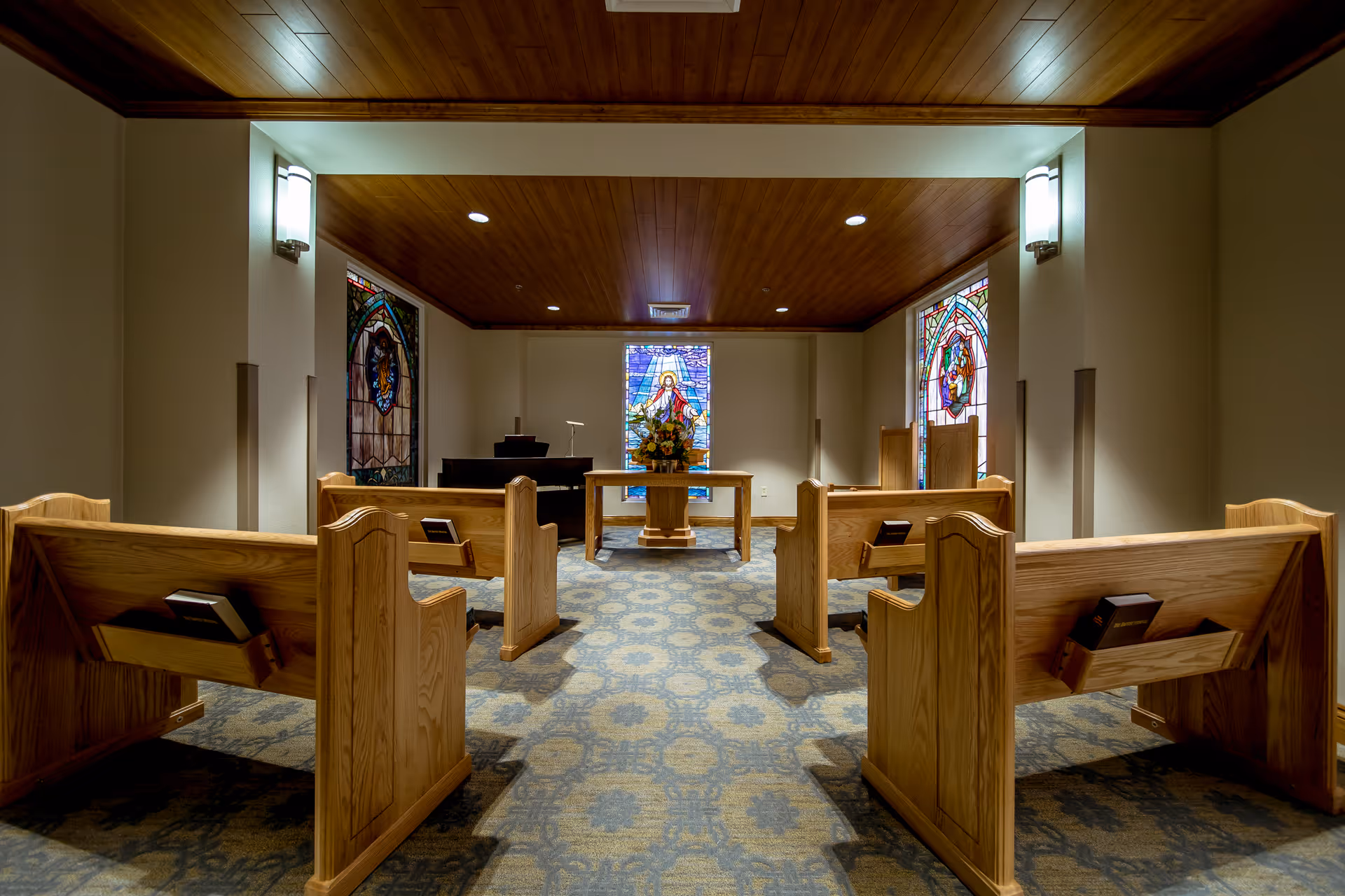 Interior view of a small chapel with wooden pews arranged in two rows facing an altar. The altar has a floral arrangement and is positioned in front of a stained glass window depicting a religious figure. Additional stained glass windows are on the side walls. The ceiling is wooden with recessed lighting, and the walls are painted a light color.