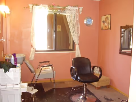 Small hair-salon room with a styling chair, shampoo sink, lace-curtained window and pink walls.