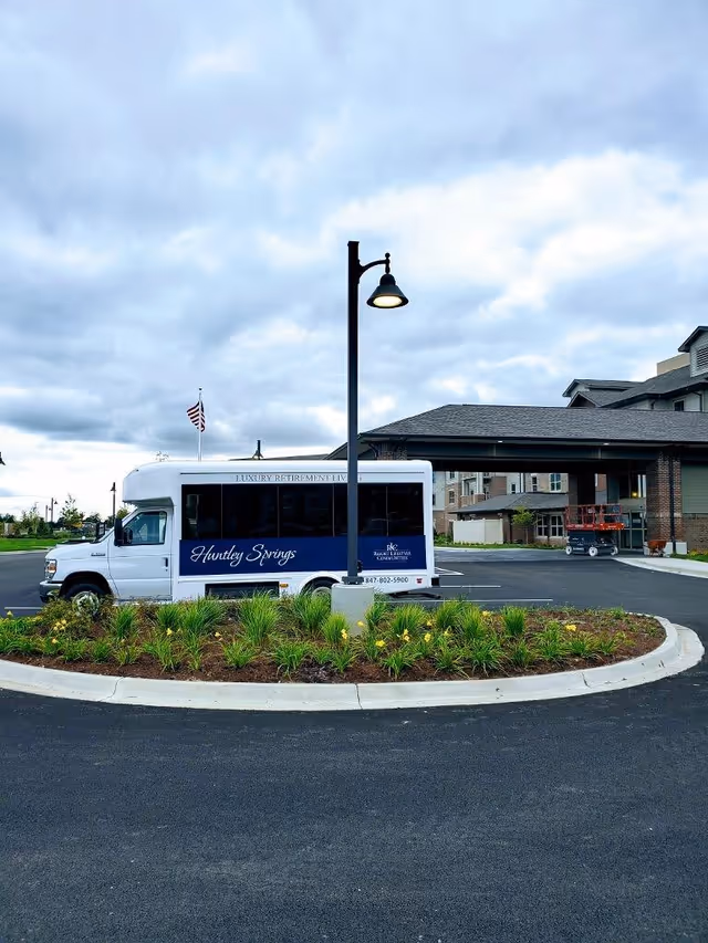 A white and blue shuttle bus with the text 'Huntley Springs' parked in front of a covered entrance to a large building under a cloudy sky. There is a landscaped roundabout with green plants and a streetlamp in the foreground.