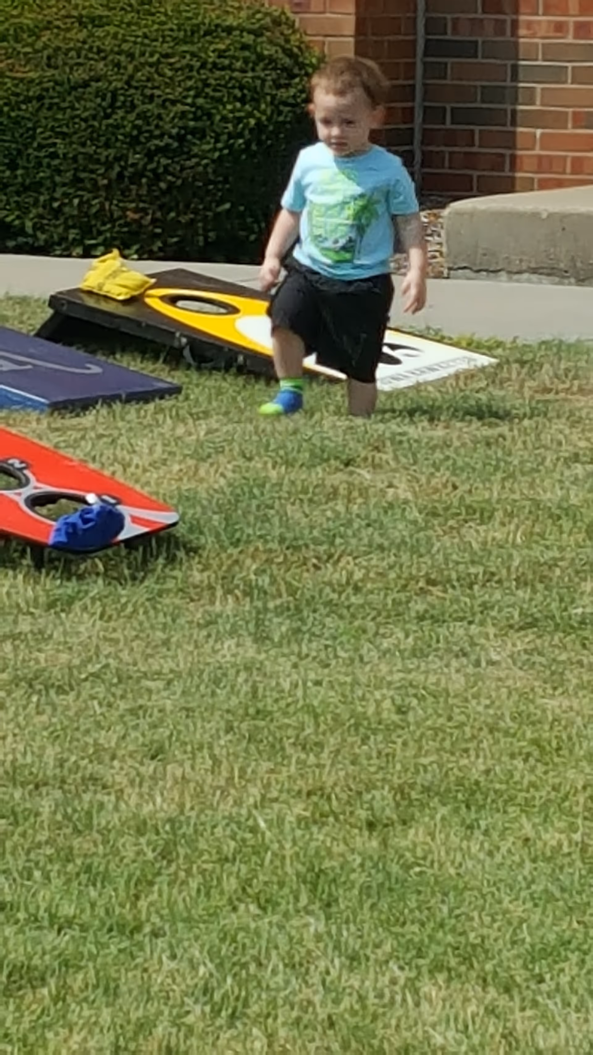 A young child walks on a grassy outdoor area with cornhole boards near a brick building.
