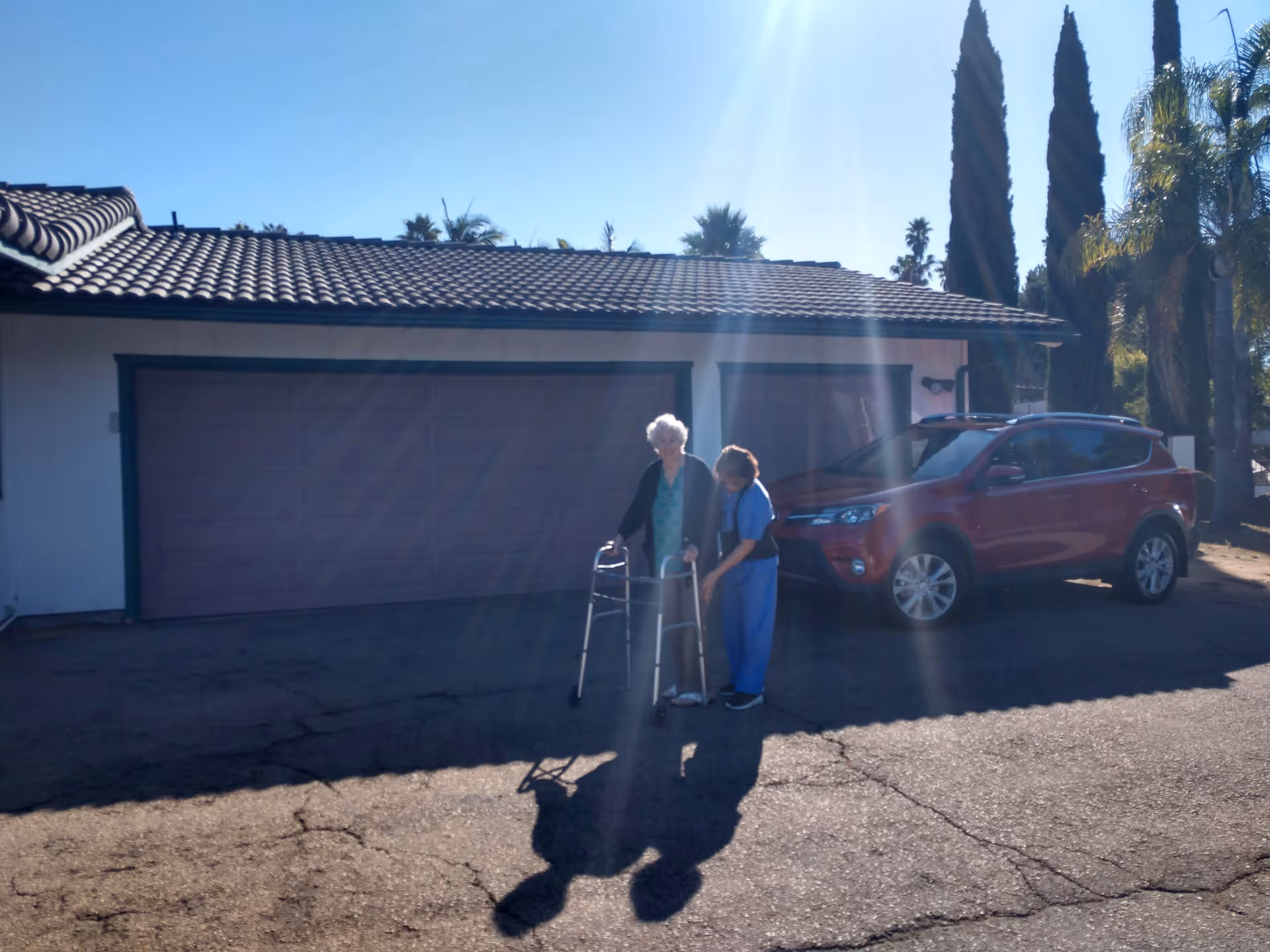 Elderly woman with walker assisted by caregiver outside Anne's Place Assisted Living, near a red SUV.