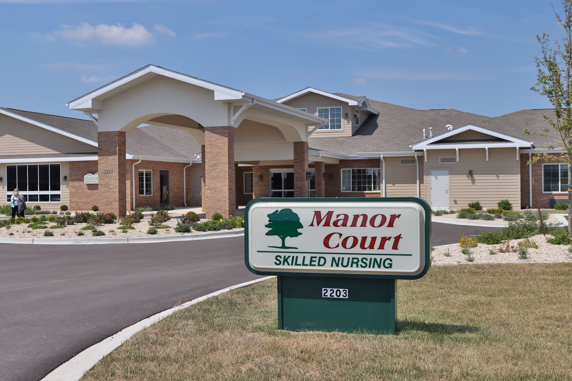 Exterior view of Manor Court Skilled Nursing facility showing the building entrance with a covered driveway and a sign in the foreground displaying the facility name and address number 2203.
