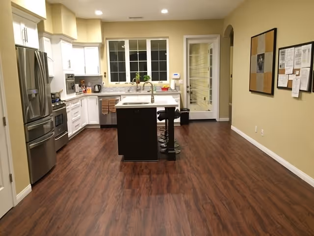 Open kitchen with stainless steel appliances, central island, bar stools, and dark wood flooring.