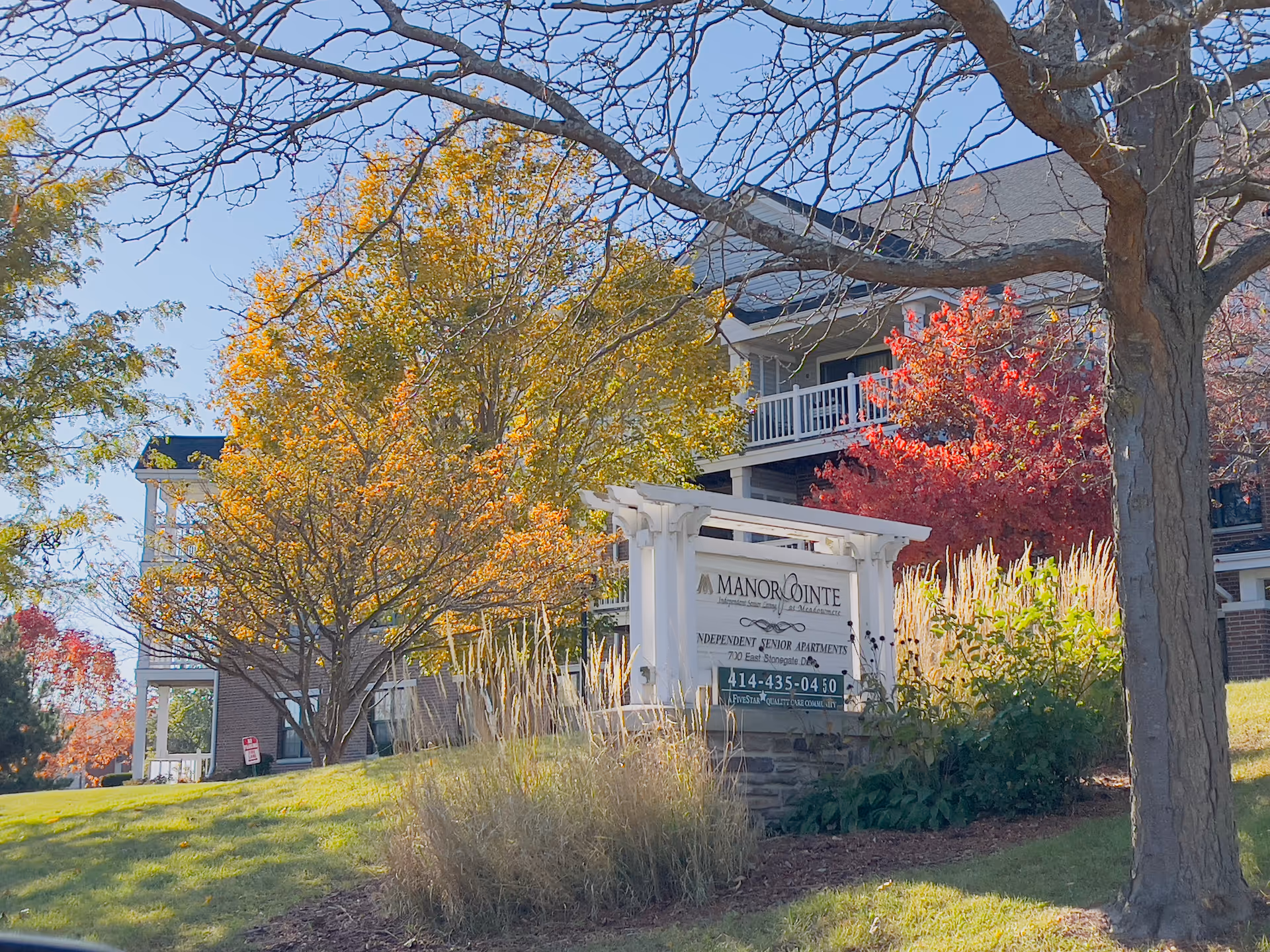 White wooden sign reading Manorpointe Independent Senior Apartments in front of a landscaped apartment building with autumn trees.