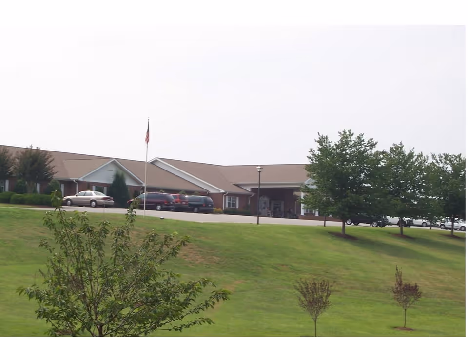 Single-story brick senior care building with a covered entrance, parked cars, a flagpole and a grassy front lawn.