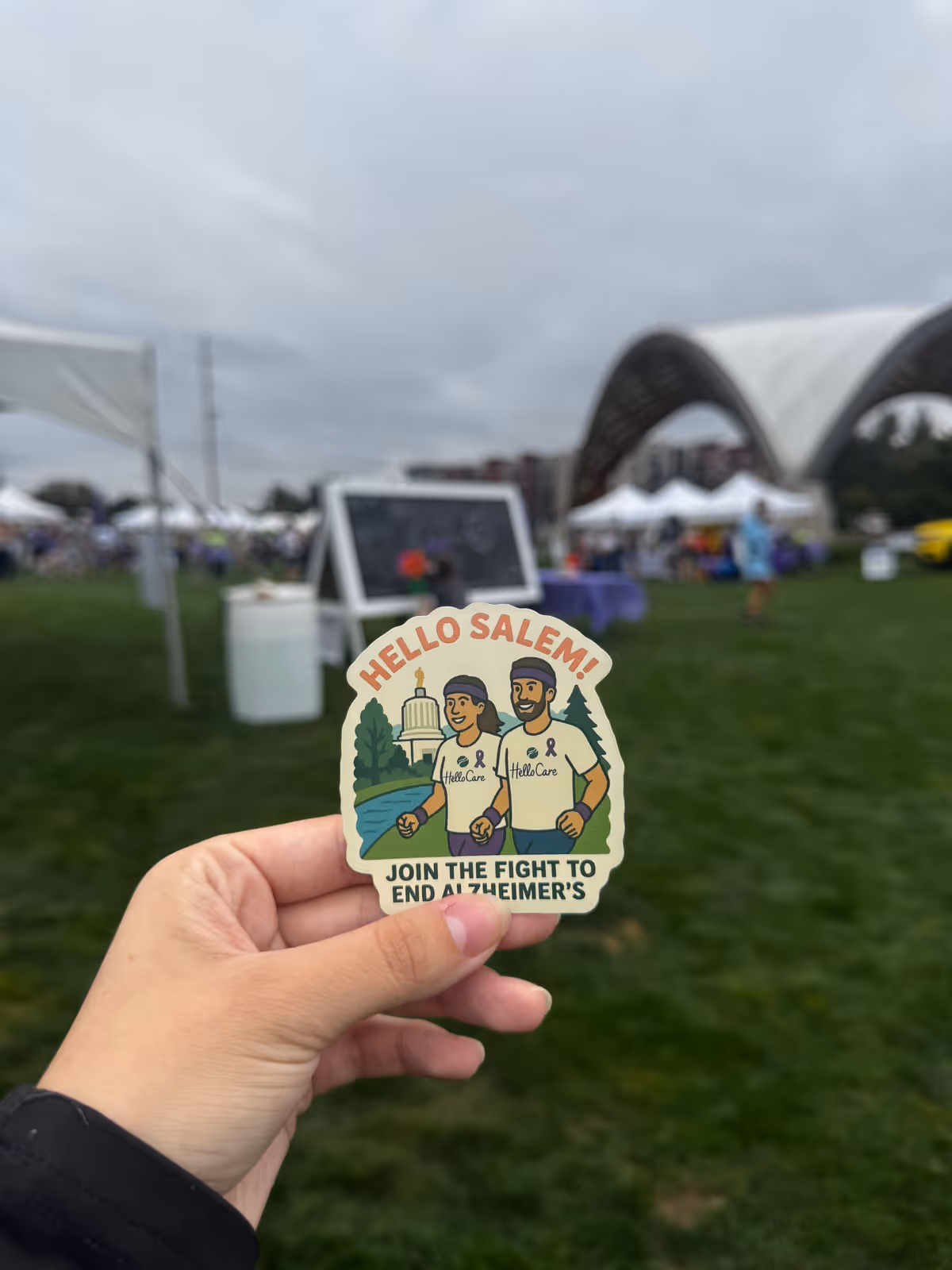 Hand holds HelloCare Salem sticker at an outdoor festival with tents