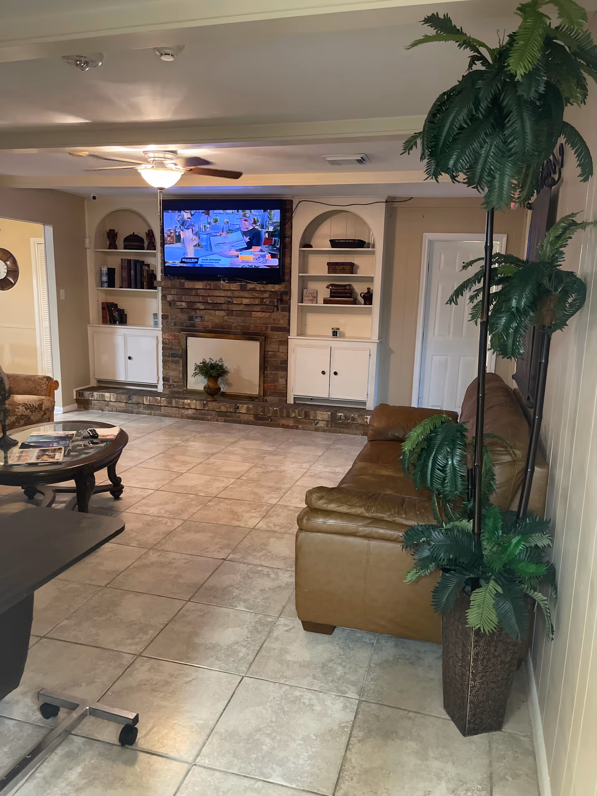 A cozy living room with a brown leather sofa, a round glass coffee table with magazines, a brick fireplace with a mounted flat-screen TV above it, built-in white shelves with books and decorative items, a ceiling fan with light, and a tall potted plant in the corner.