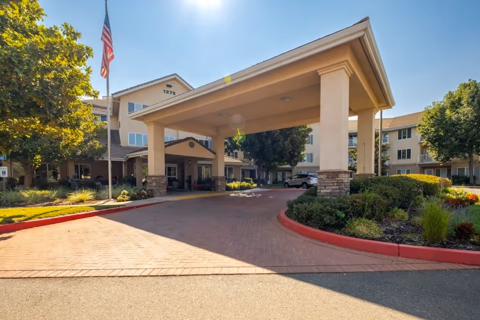 Senior living facility entrance with a wide porte-cochere, flag, and landscaped circle driveway.