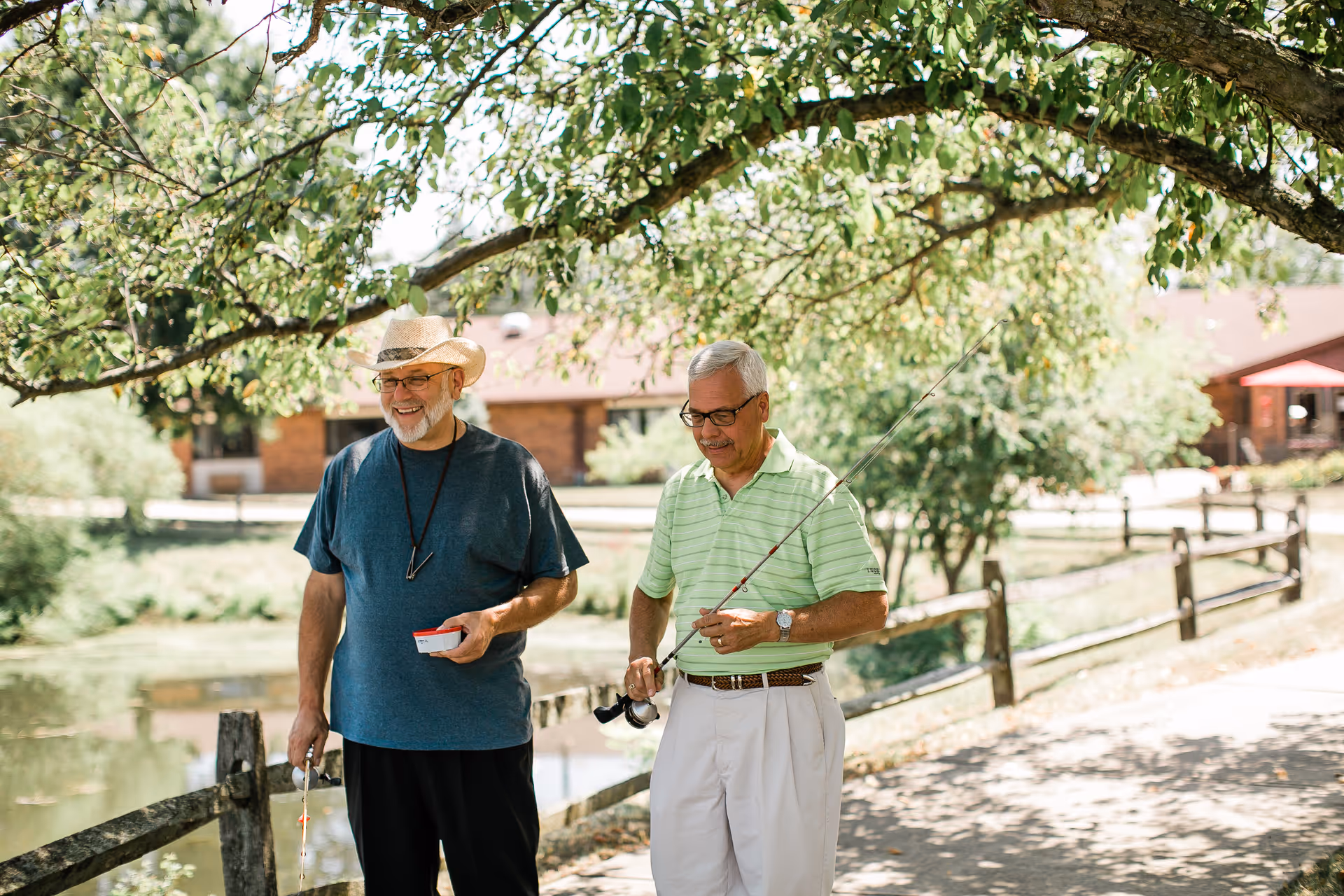 Two elderly men standing outdoors near a wooden fence by a pond, one wearing a straw hat and blue shirt holding fishing gear, the other in a green striped polo shirt holding a fishing rod, with trees and buildings in the background.
