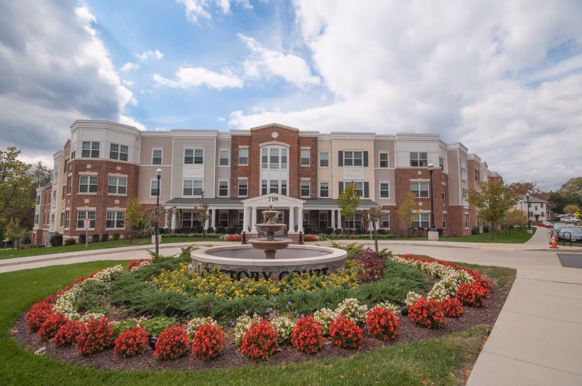Front view of a brick senior apartment building with a circular fountain and colorful flower beds