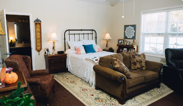 A cozy bedroom in a senior living facility featuring a bed, couches, lamps, and a rug.