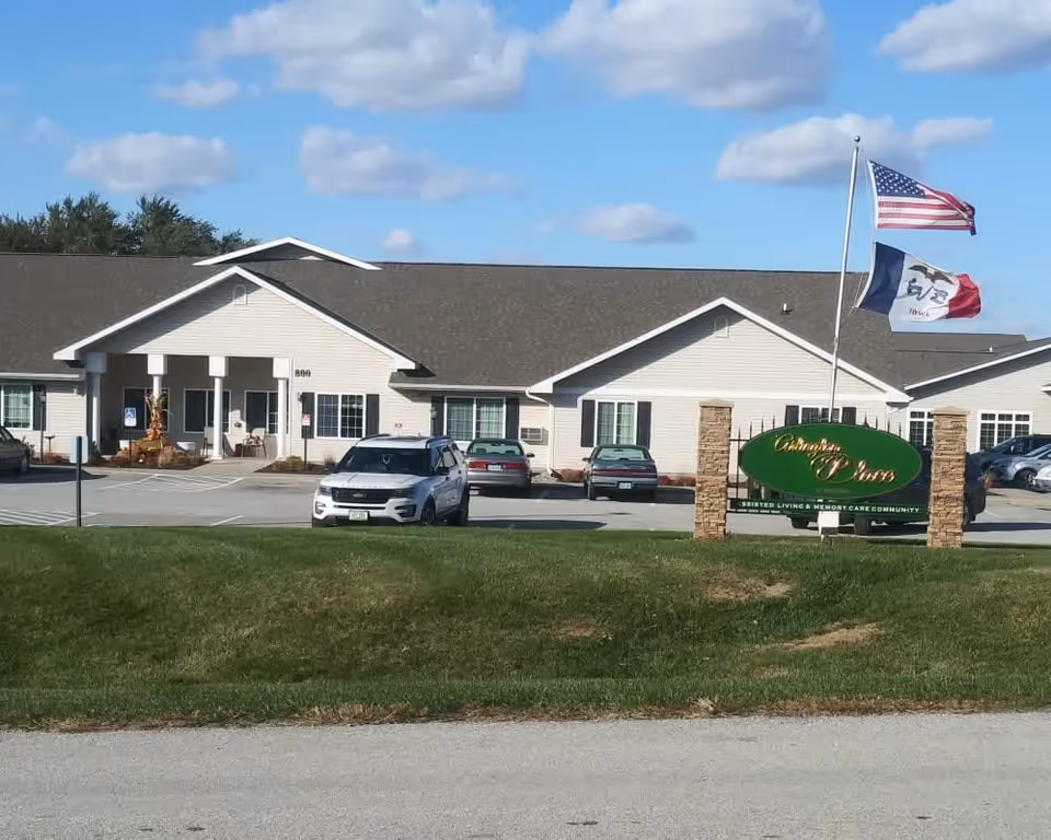 Exterior view of Arlington Place at Red Oak, a single-story assisted living and memory care community building with a parking lot in front. Two flags, the American flag and the Iowa state flag, are flying on a flagpole near a green sign with the facility's name. The sky is partly cloudy.
