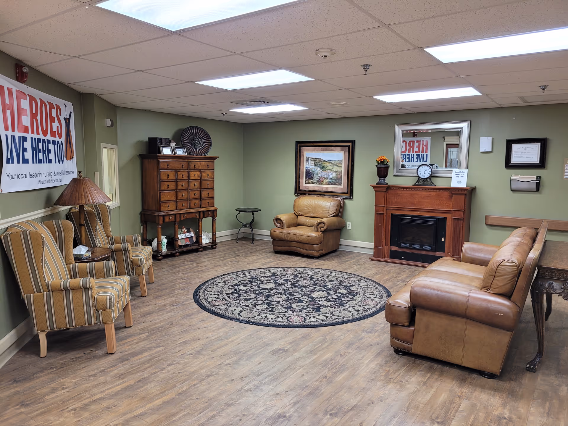 A cozy common room in a senior living facility with leather sofas and armchairs around a round rug and a fireplace, a cabinet and a banner on the wall.
