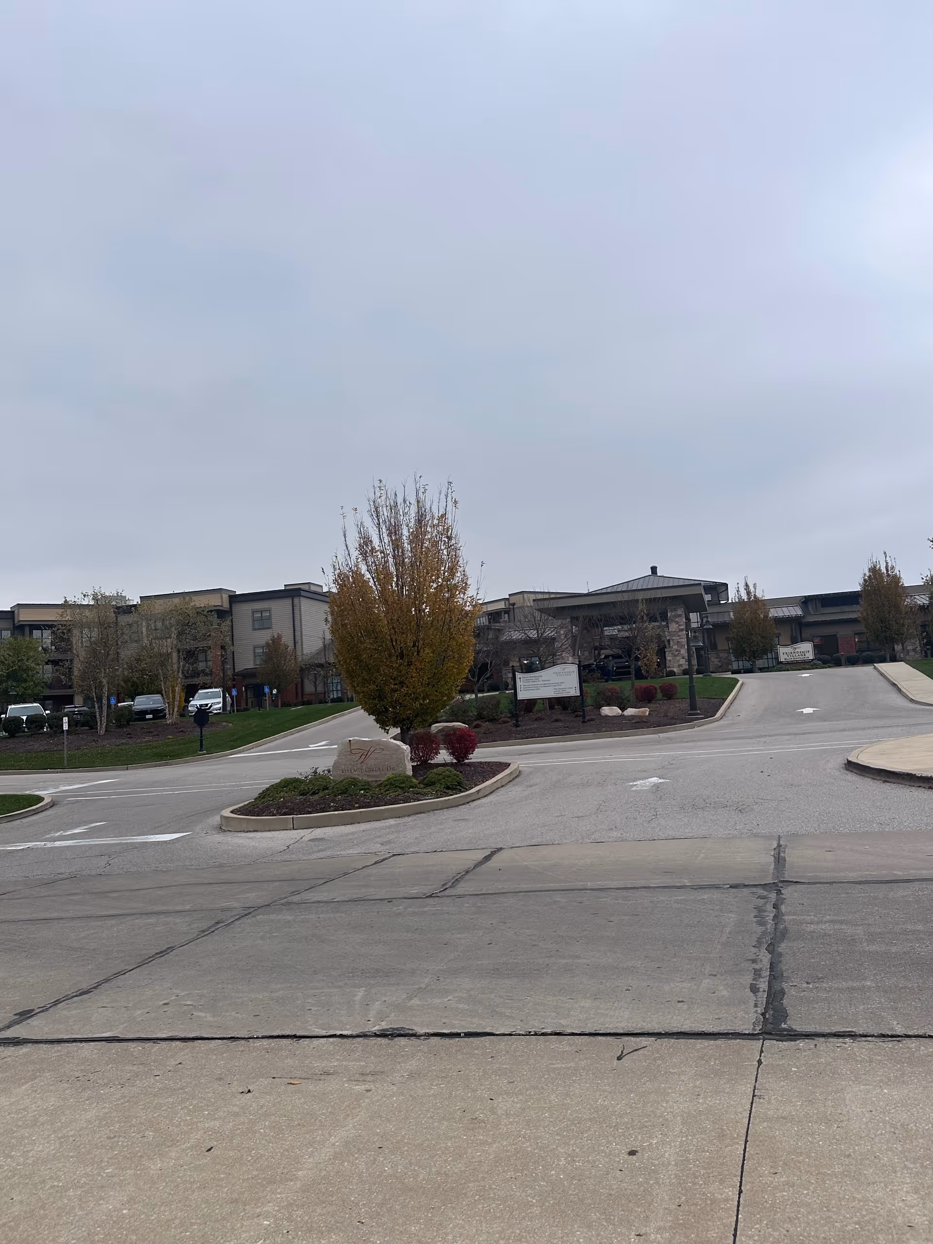 Exterior view of Friendship Village Sunset Hills senior living facility showing the driveway entrance, landscaped area with a tree and bushes, and the multi-story building under a cloudy sky.