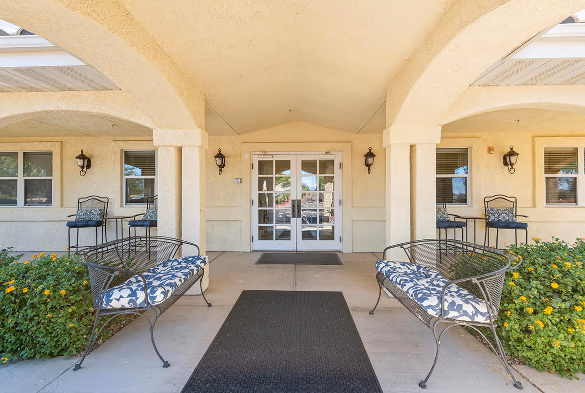 Entrance area of a senior living facility with two cushioned metal benches facing each other, two small tables with chairs against the wall, beige stucco walls, and double glass doors under an arched covered walkway.
