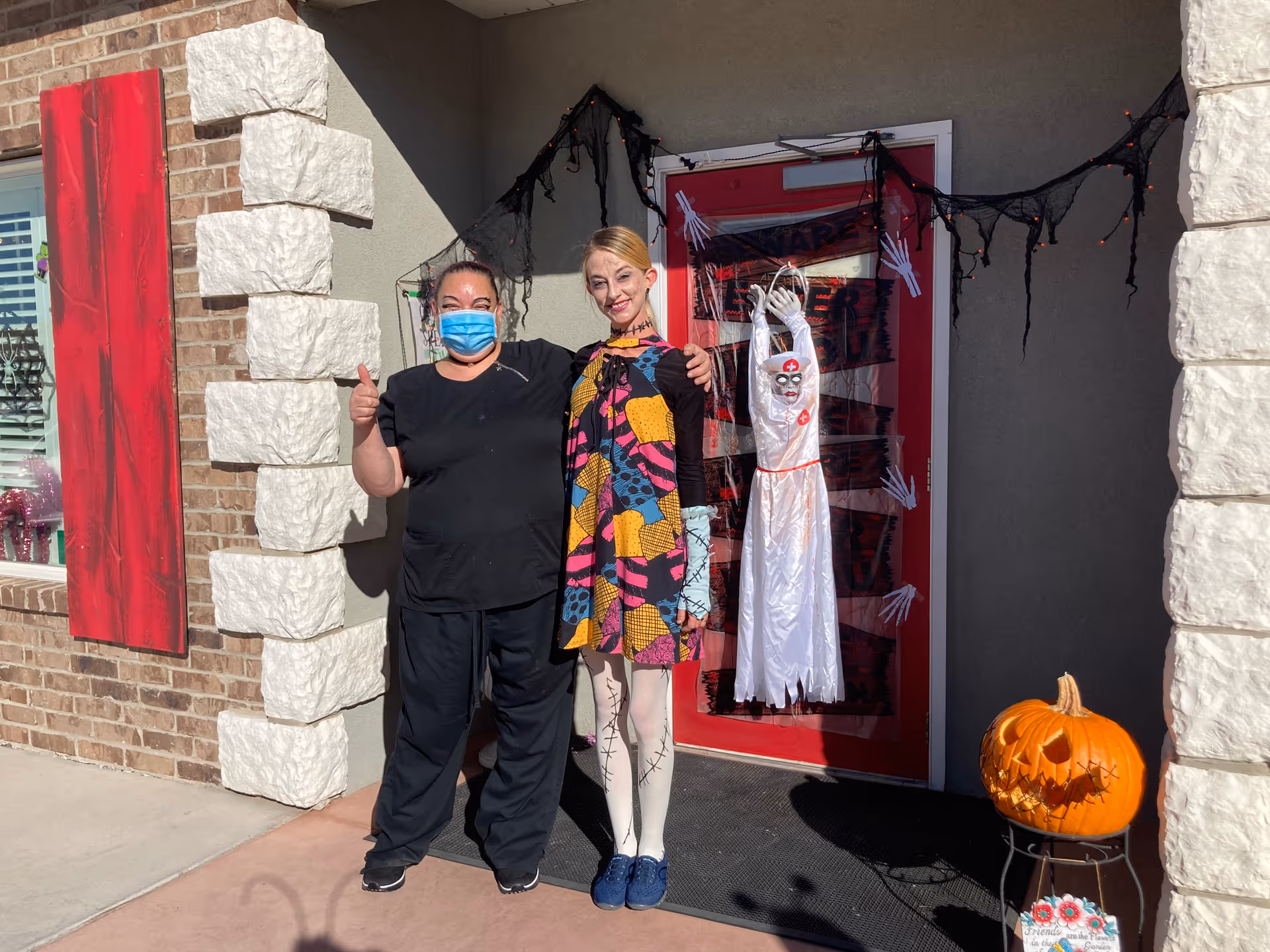 Two women standing outside the entrance of a building decorated for Halloween. One woman is wearing a black outfit and a blue face mask, giving a thumbs up. The other woman is dressed in a colorful Halloween costume with face paint. The red door behind them is decorated with black spider webs, skeleton hands, and a hanging ghost figure. A carved pumpkin with a spooky face is placed on a stand to the right of the door.
