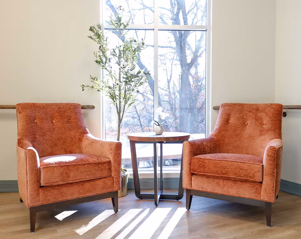 Two orange upholstered chairs flank a small table by a large window in a bright interior.