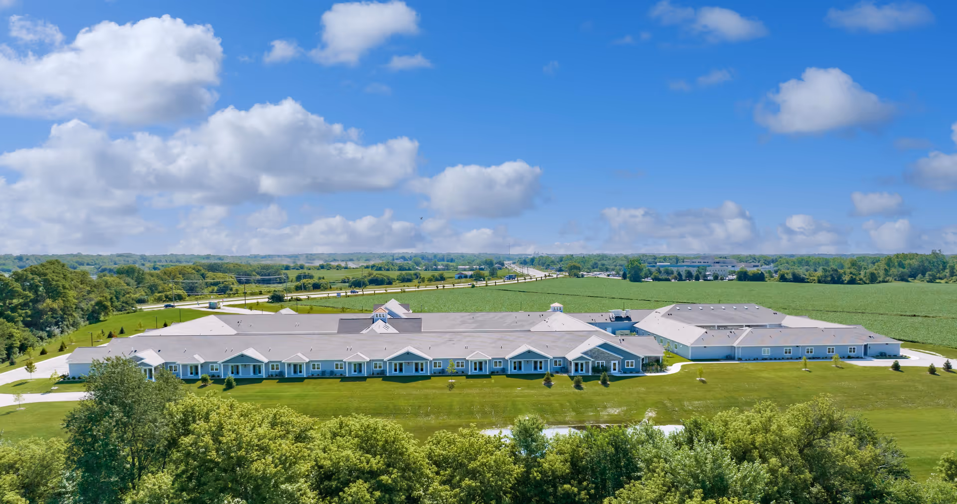 Aerial view of a long single-story senior living building set in green lawns and surrounding farmland under a blue sky with clouds.