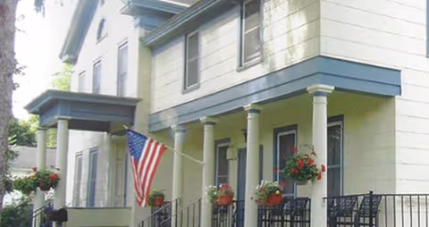 Exterior view of a yellow senior living facility with a porch, columns, and an American flag.