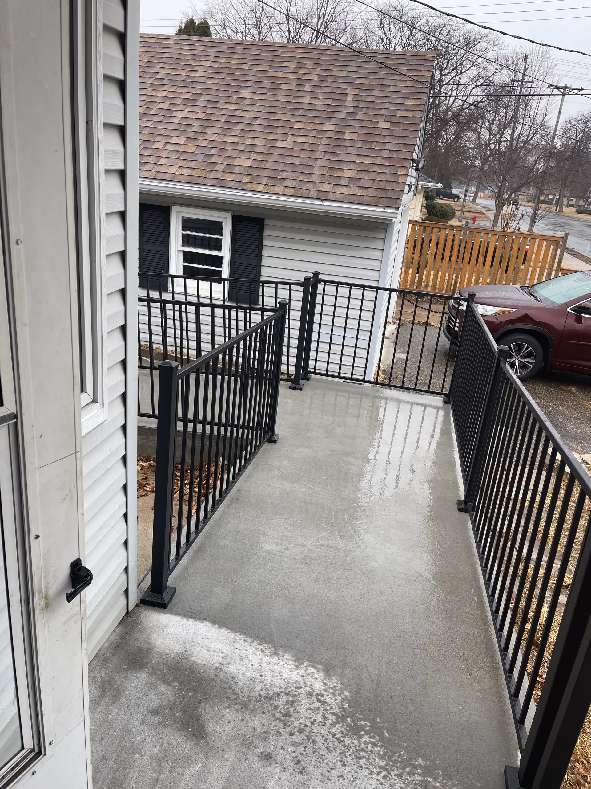 A wet concrete balcony with black metal railing along the side of a light siding building.
