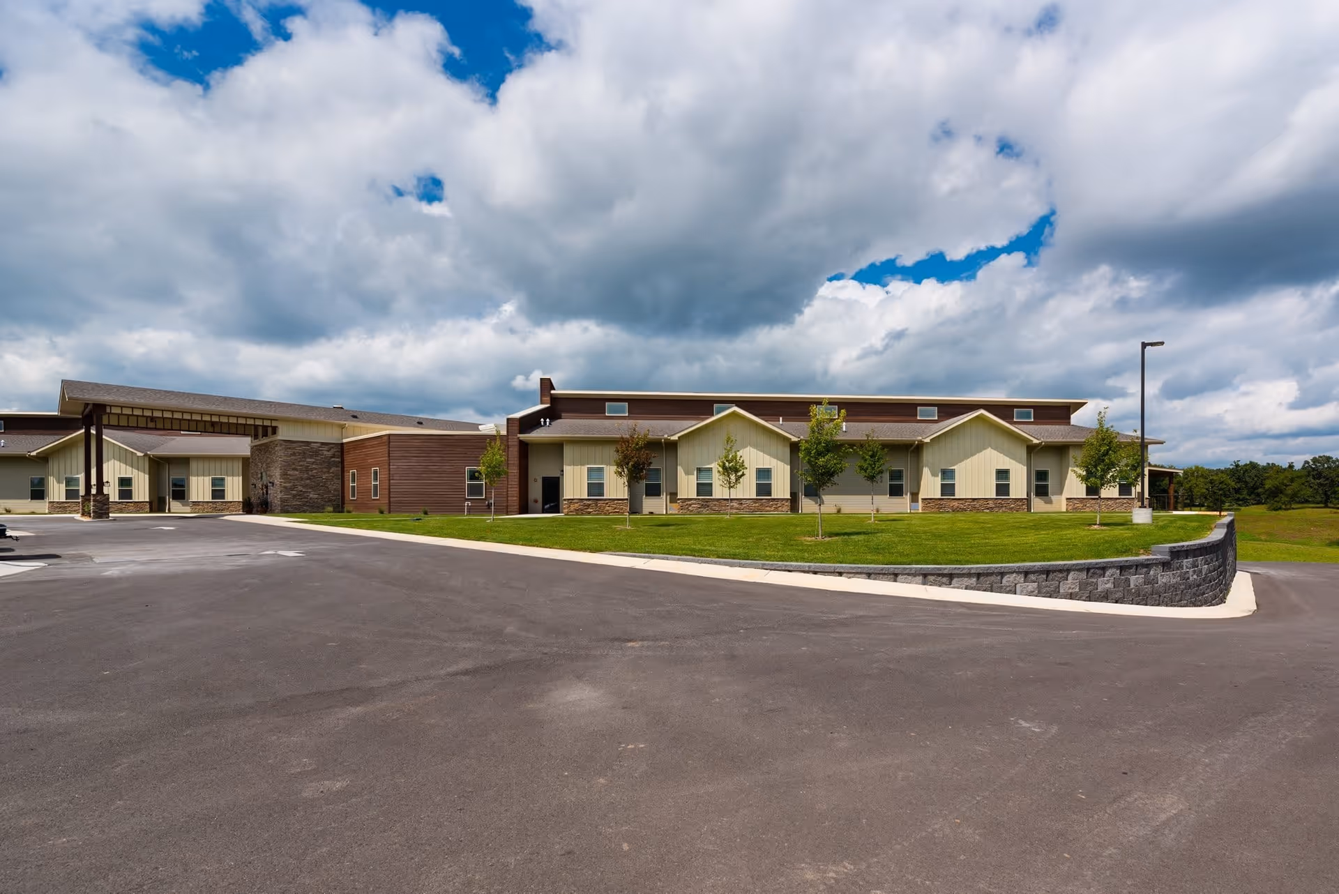 Exterior view of Webwood Assisted Living facility showing a single-story building with beige and brown siding, a stone accent wall, a covered entrance, green lawn with young trees, and a paved driveway under a partly cloudy sky.