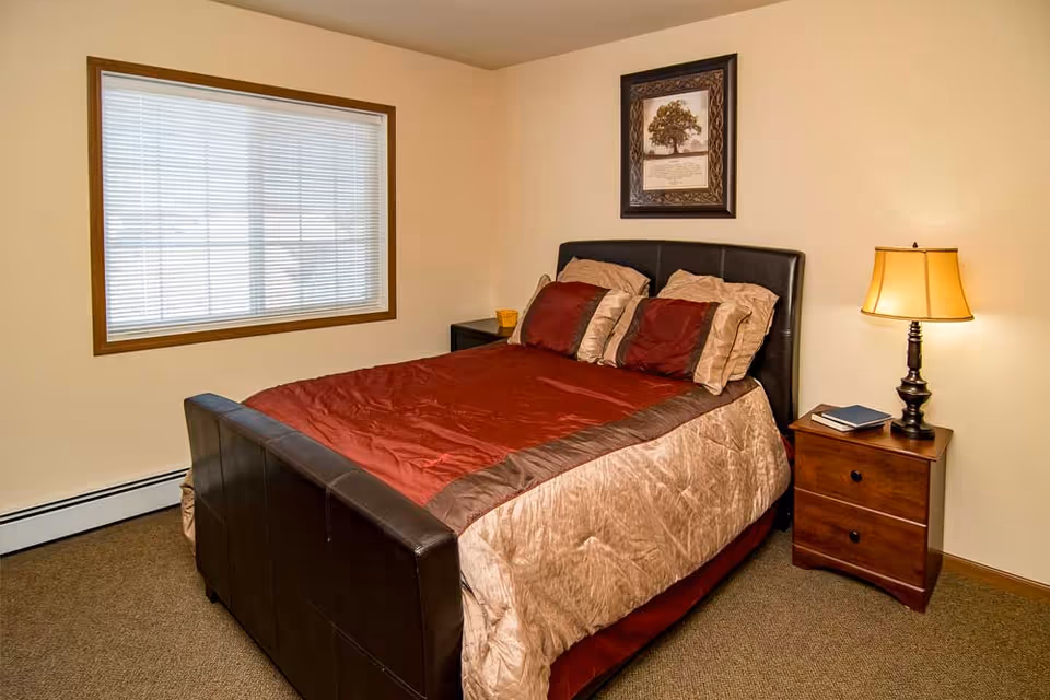 Simple bedroom with a bed dressed in red and beige bedding, a wooden nightstand with a lamp, a window with blinds, and framed artwork on the wall.