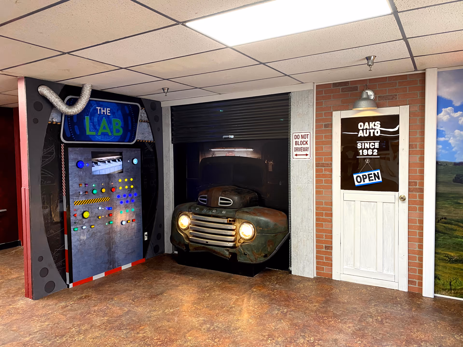 An indoor scene featuring a decorative setup with a vintage car front partially inside a black garage door, a white door with a sign reading 'Oaks Auto Since 1962 OPEN,' and a colorful control panel labeled 'The Lab' with various buttons and lights on the left side.
