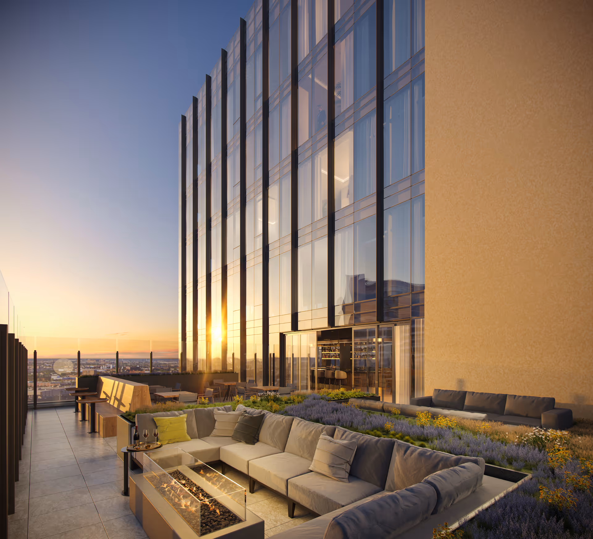A modern outdoor rooftop lounge area at sunset with a large L-shaped cushioned sofa surrounding a rectangular fire pit. The space includes additional seating, plants, and a glass railing offering a city view. The building has tall glass windows reflecting the sunset light.