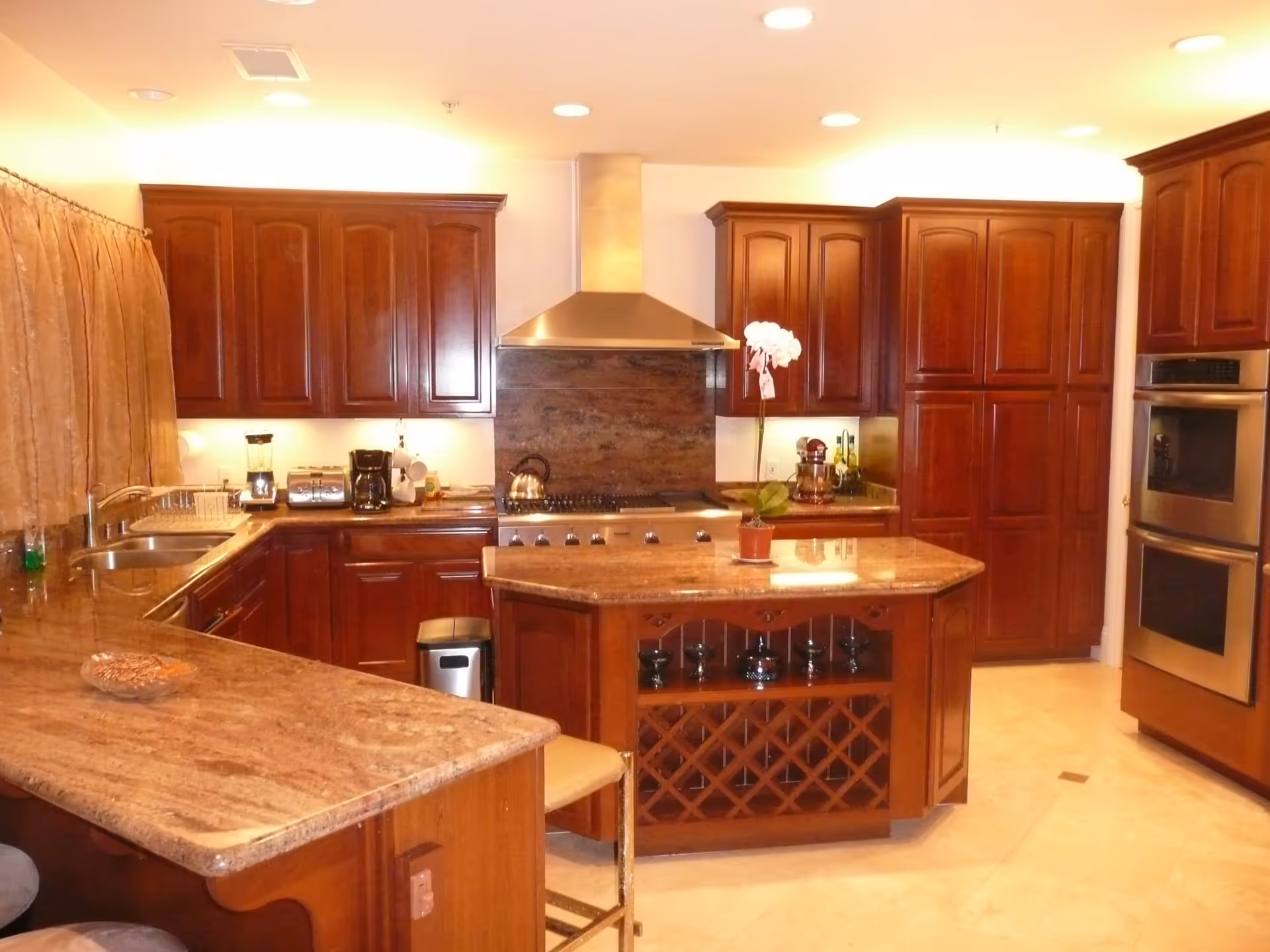 A spacious kitchen with wooden cabinets and granite countertops. The kitchen features a central island with a built-in wine rack, a stainless steel range hood above a stove, double wall ovens, a coffee maker, toaster, and a sink under a window with curtains. There is a potted orchid on the island and a beige tiled floor.