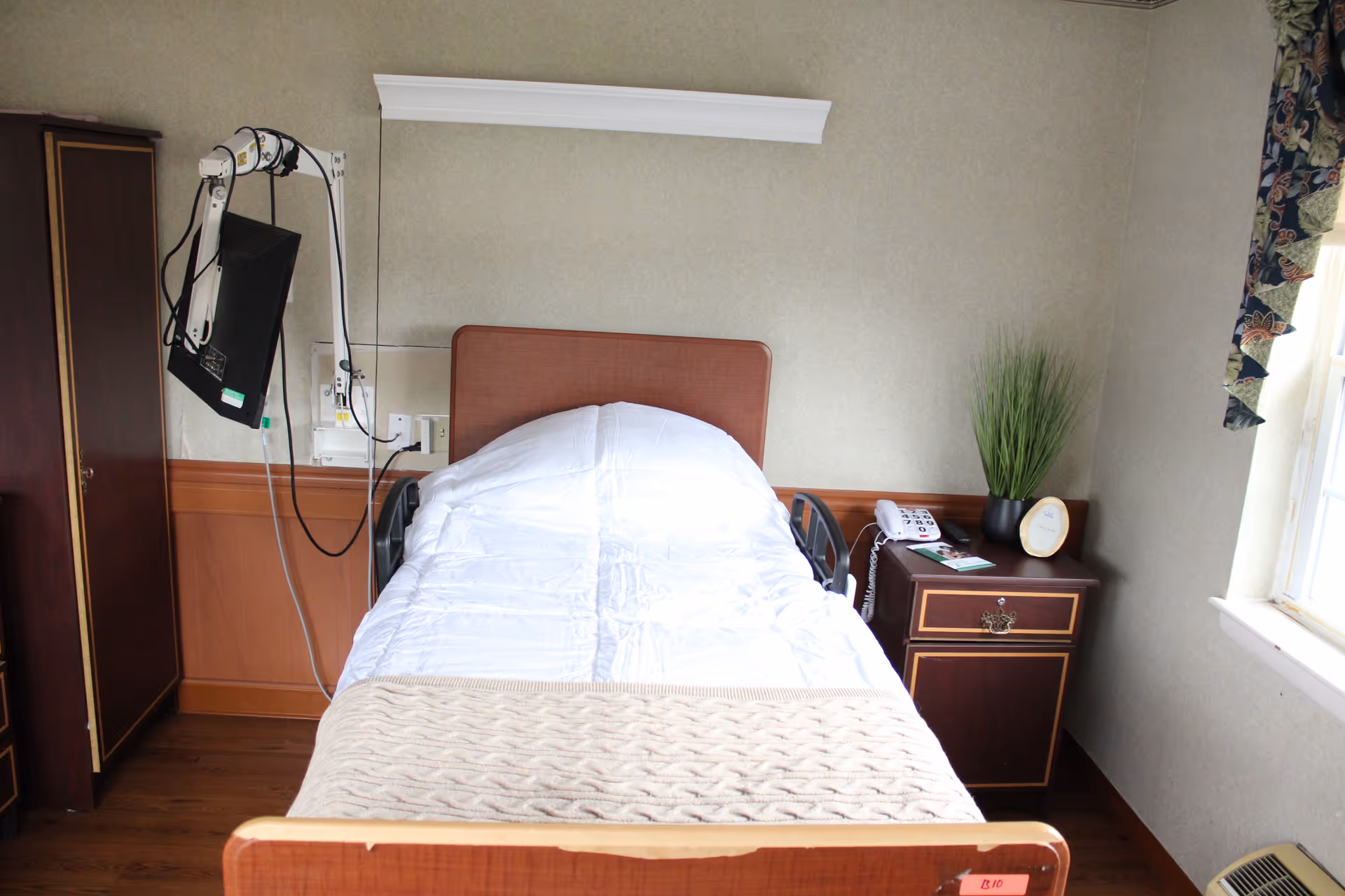 A single hospital-style patient bed with white bedding, medical monitor, and nightstand in a modest room.