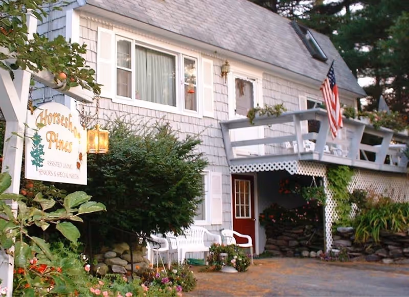 Front of a two-story gray shingled building with a porch, American flag, and a 'Horseshoe Pines' sign surrounded by shrubs and flowers.