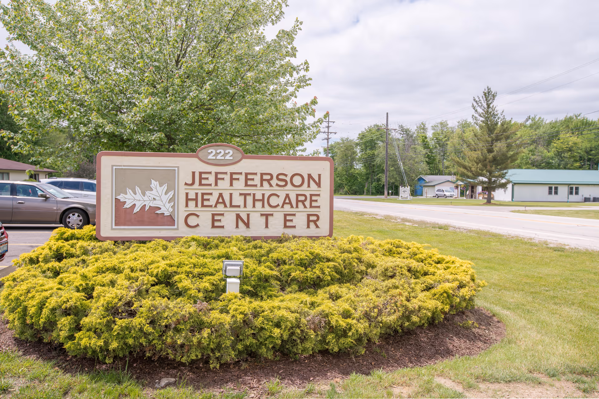 Outdoor view of a sign for Jefferson Healthcare Center with the address number 222. The sign is surrounded by green shrubs and is located near a road with trees and buildings in the background under a cloudy sky.