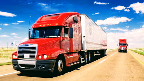 Red semi-trucks driving on a wide highway beneath a bright blue sky.