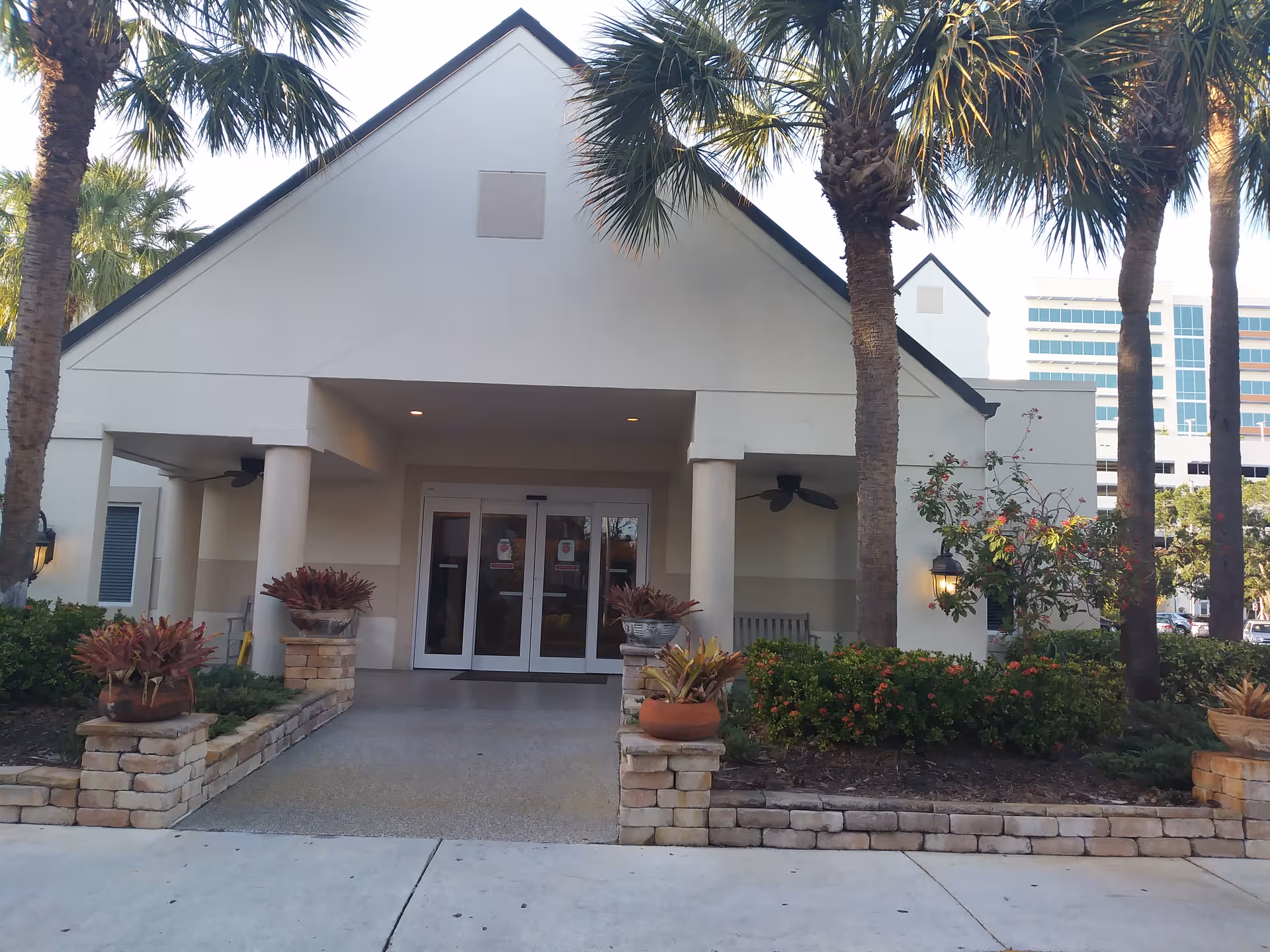 White building entrance with columns, glass doors, and palm trees along a landscaped walkway.