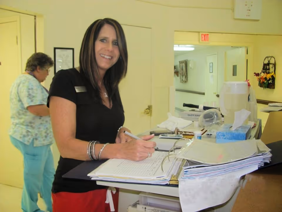 A woman with long dark hair wearing a black shirt and red pants is smiling and writing in a large binder on a counter in a nursing facility. In the background, another woman in medical scrubs is walking past a yellow door. The setting appears to be a hallway or nurse's station with medical supplies and paperwork on the counter.
