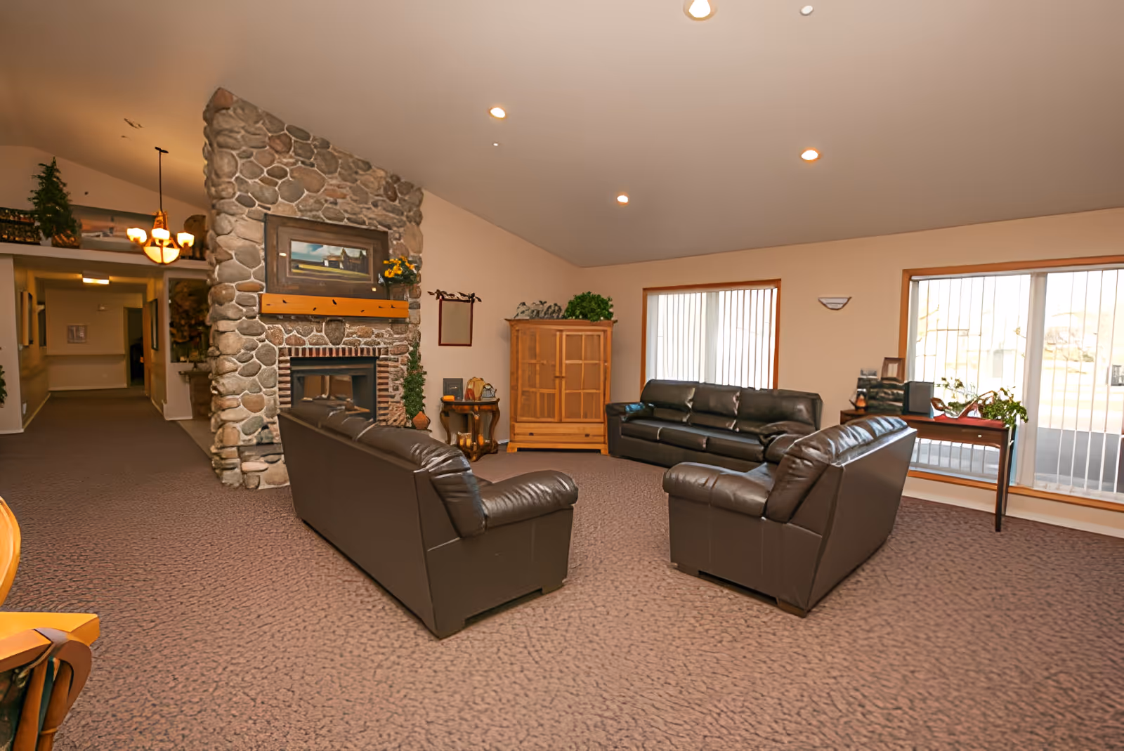 A cozy living room with three dark leather sofas arranged around a stone fireplace. The fireplace has a wooden mantle with a painting and decorative items on top. The room has beige walls, carpeted floor, large windows with vertical blinds, and wooden furniture including a cabinet and a small table with plants.