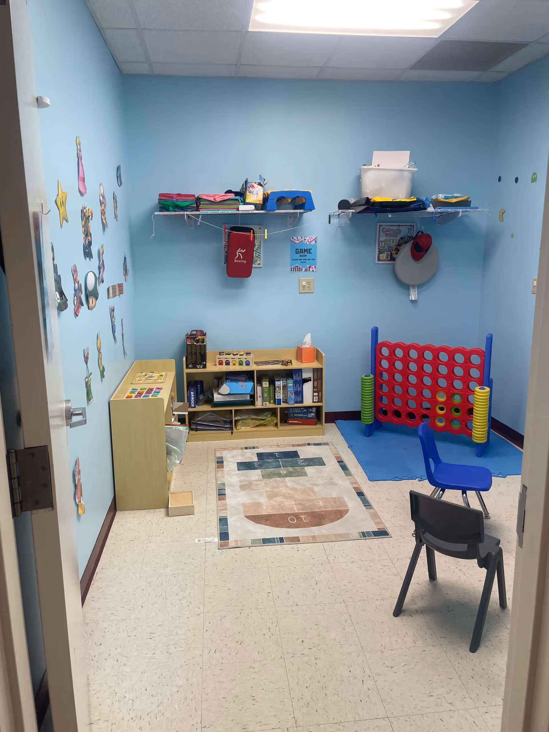Bright blue therapy room with shelves, mats, chairs, and a large Connect Four game.