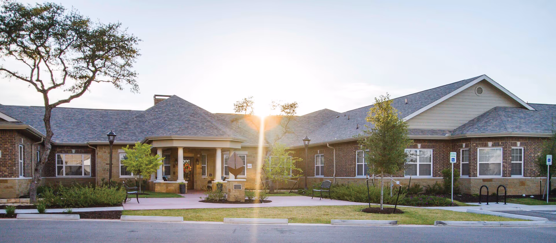 Sunlit brick senior living facility exterior with entrance, landscaping, and benches.