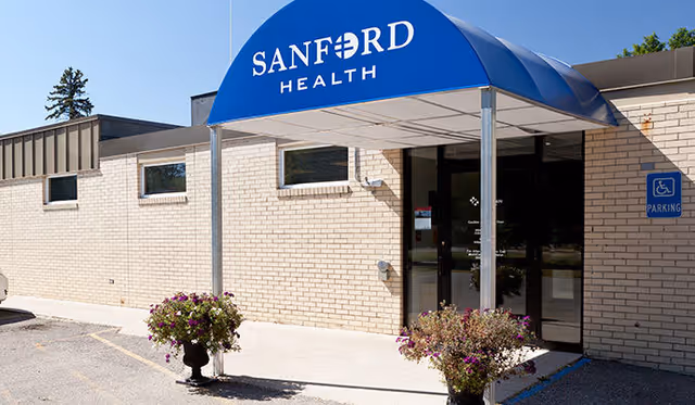 Brick facility entrance with blue Sanford Health canopy and potted flowers.